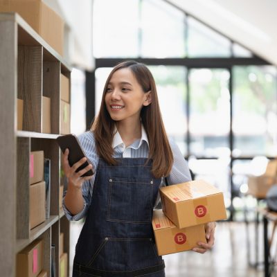 Attractive asian woman hold package box, using mobile phone checking stock order, startup SME