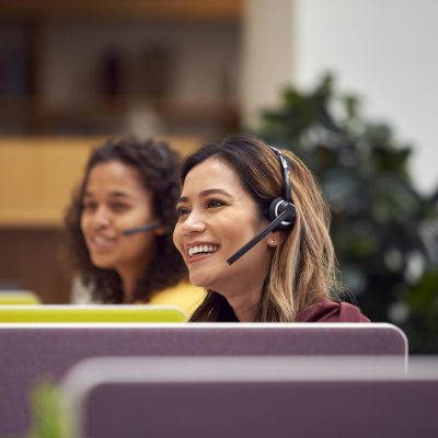 Mature Businesswoman Wearing Phone Headset Talking To Caller In Busy Customer Services Centre